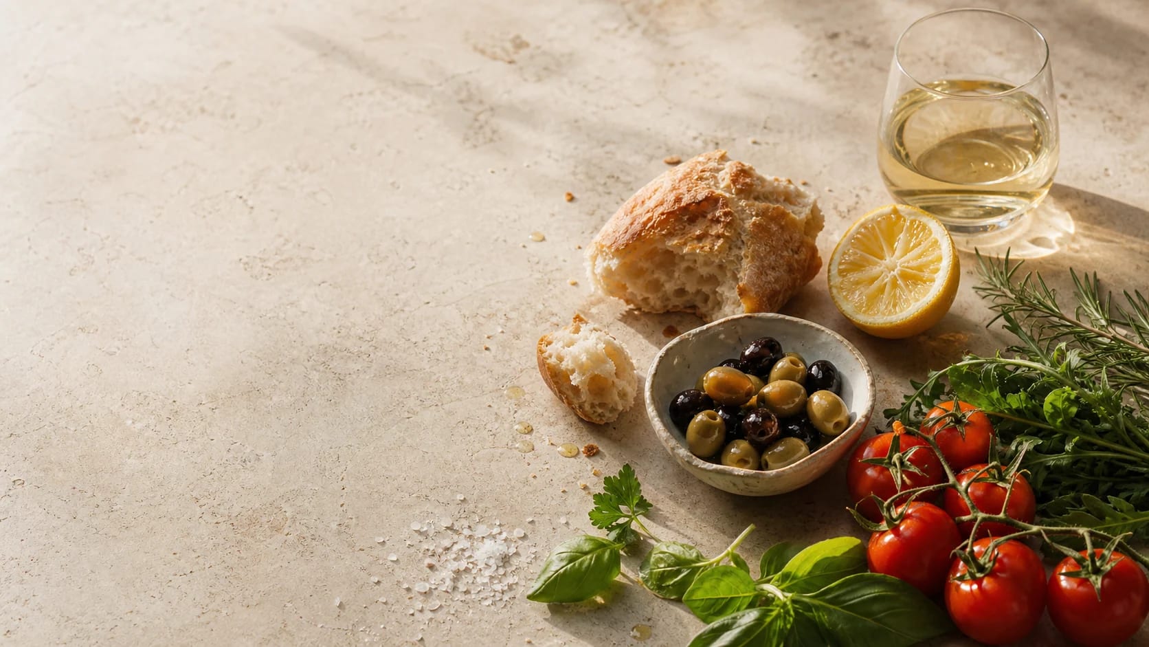 Bread, olives, tomatoes, lemon and fresh herbs arranged on pale stone.