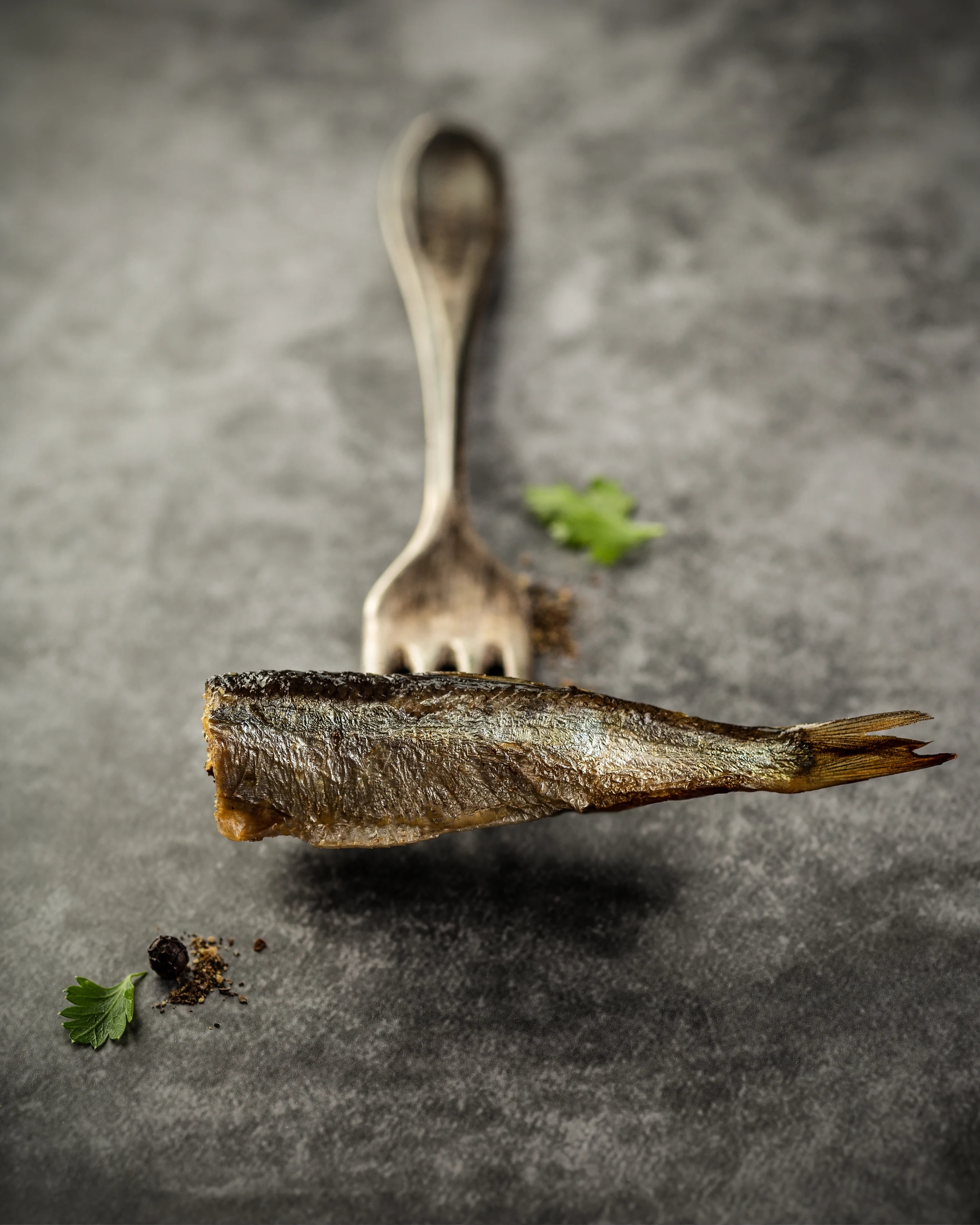 A smoked sprat presented above a dark stone surface with a fork and fresh herbs