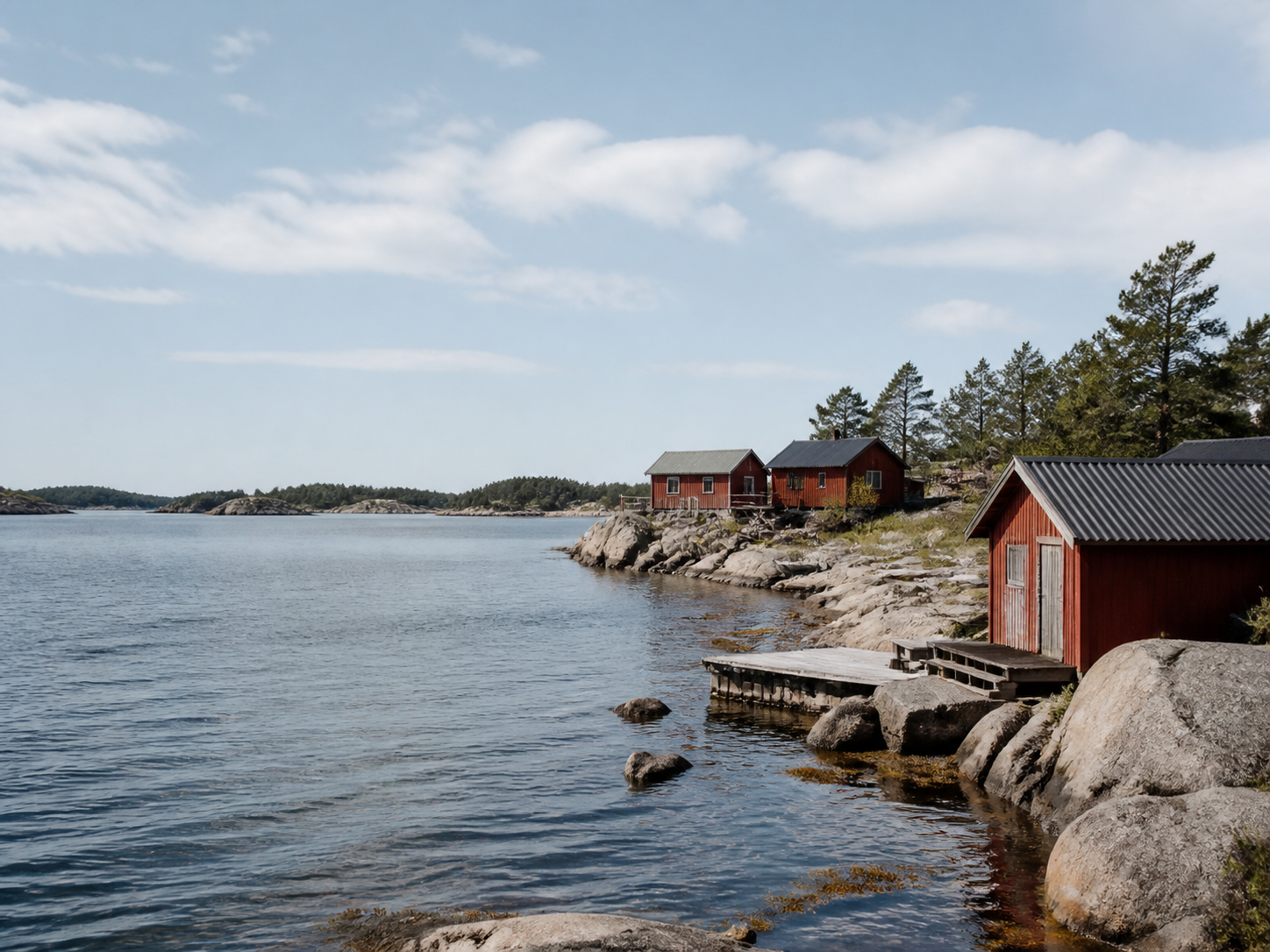 A bright Baltic archipelago coast with low rocks, calm water and weathered red boathouses.